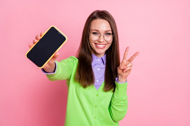 Il ritratto dello smartphone positivo della tenuta della ragazza fa il v-sign isolato sopra il fondo di colore pastello