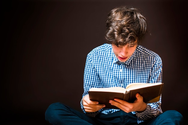 Il ritratto dello studente adolescente si siede sul pavimento e legge un libro su fondo scuro isolato b