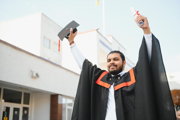 Il ritratto dello studente laureato indiano bello in bagliore di laurea con il diploma