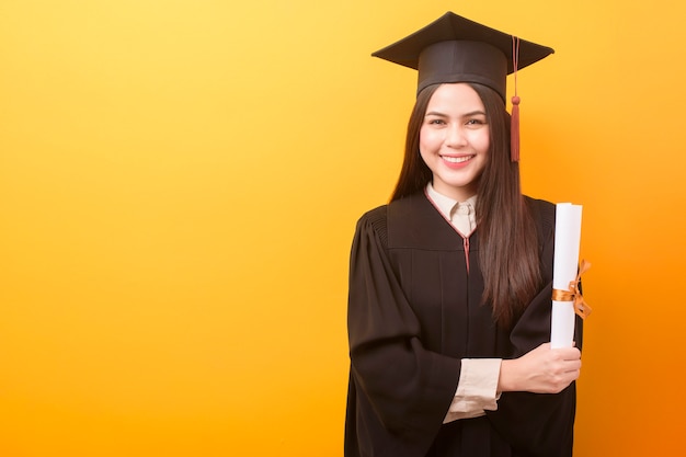 Il ritratto di bella donna felice in abito di graduazione sta tenendo il certificato di istruzione su fondo giallo