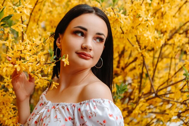 Il ritratto di bella donna tenera ha vestito l'abito fiorito rosa che posa vicino all'albero giallo della forsizia