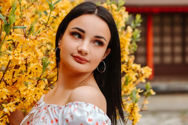 Il ritratto di bella donna tenera ha vestito l'abito fiorito rosa che posa vicino all'albero giallo della forsizia