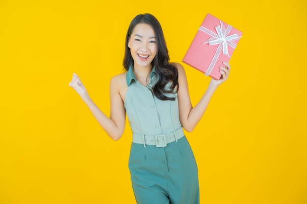Il ritratto di bella giovane donna asiatica sorride con il contenitore di regalo rosso sulla parete gialla
