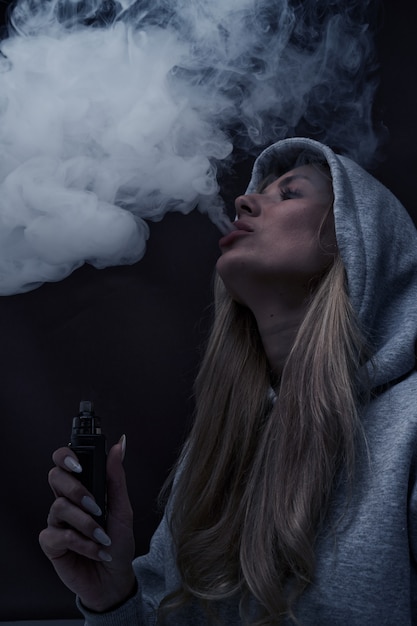 Il ritratto di bella ragazza bionda in felpa con cappuccio grigia fuma vape isolato su sfondo nero studio, nuvola di fumo di vapore, mini narghilè