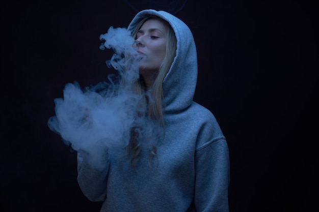 Il ritratto di bella ragazza bionda in felpa con cappuccio grigia fuma vape isolato su sfondo nero studio, nuvola di fumo di vapore, mini narghilè