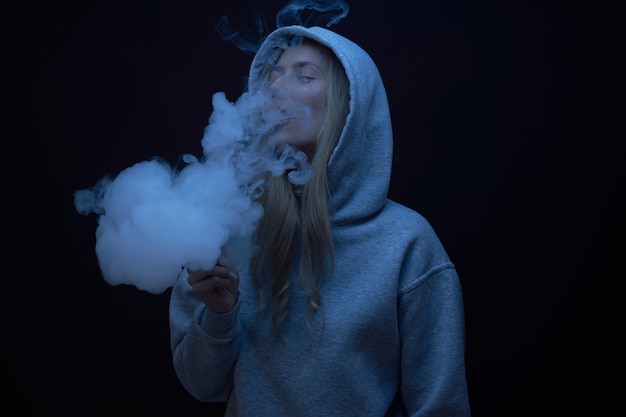 Il ritratto di bella ragazza bionda in felpa con cappuccio grigia fuma vape isolato su sfondo nero studio, nuvola di fumo di vapore, mini narghilè