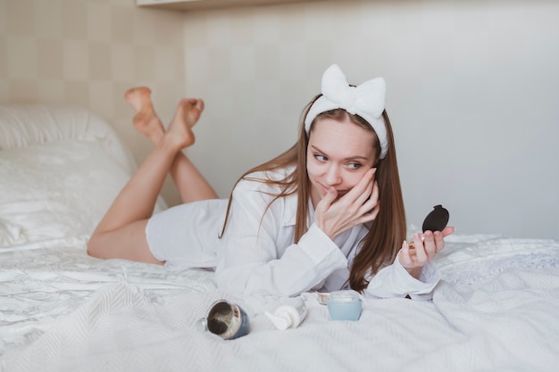 Il ritratto di bella ragazza con capelli lunghi a letto si prende cura della sua pelle. Utilizzo di creme cosmetiche