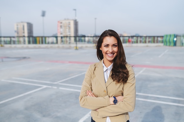 Il ritratto di bello brunette caucasico si è vestito in rivestimento beige che si leva in piedi sul parcheggio con le braccia attraversate.