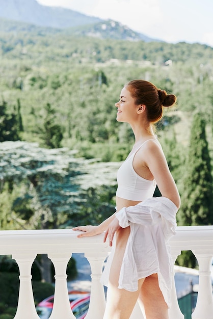 Il ritratto di giovane bella donna in una camicia bianca ammira la natura verde sulla giornata di sole del balcone