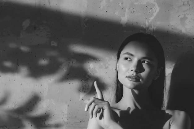 Il ritratto di giovane donna allegra sveglia mostra il dito e distoglie lo sguardo sul vecchio fondo del muro con ombra dalla foglia. Concetto pubblicitario di stile di vita sano e cura di sé. Copia spazio per sito o SPA