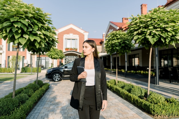 Il ritratto di giovane donna di affari abbastanza concentrata caucasica che cammina con la compressa digitale contro l'automobile nera e l'ufficio moderno concentrano all'aperto