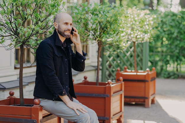 Il ritratto di un allegro maschio barbuto di mezza età parla con qualcuno tramite il cellulare ha un aspetto accattivante trascorre il tempo libero in un bellissimo giardino o parco gode di un'atmosfera calma e di un bel paesaggio