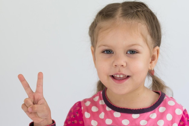 Il ritratto di un bambino caucasico di tre anni mostra le dita per mostrare il numero due o firmare la vittoria guardando la fotocamera sullo sfondo bianco