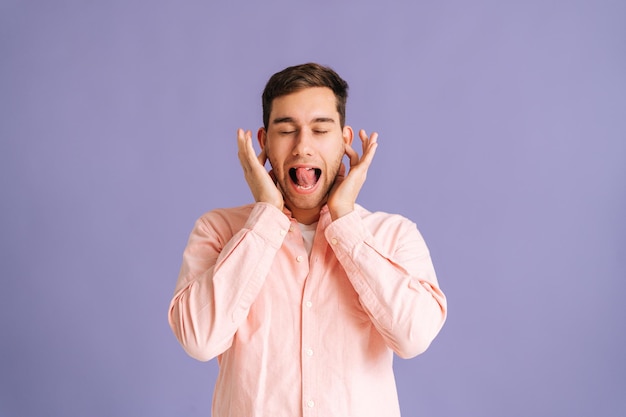 Il ritratto di un giovane infastidito che copre le orecchie con le mani dice bla bla non voglio ascoltare su sfondo rosa isolato in studio. Maschio bello annoiato che mostra contenuti senza senso.