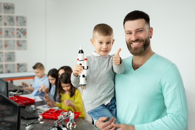 Il ritratto di un giovane padre con suo figlio in braccio mostra i pollici in su in una lezione di robotica