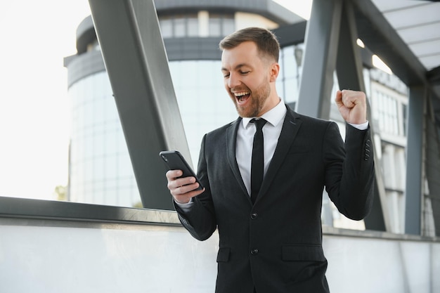 Il ritratto di un giovane uomo d'affari eccitato si è vestito con il telefono cellulare della tenuta del vestito che celebra