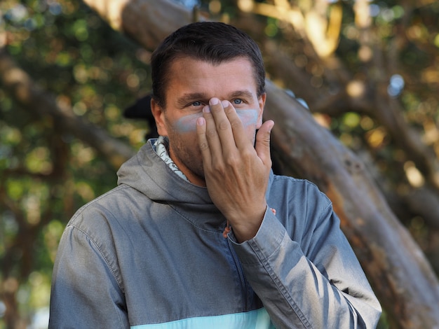 Il ritratto di un surfista maschio si è vestito con una felpa con cappuccio che applica la protezione solare colorata sul suo fronte. Ambiente tropicale.