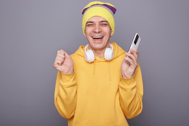 Il ritratto di un uomo hipster estremamente felice che indossa abiti casual in piedi contro il muro grigio che tiene il telefono cellulare e urla di felicità ha un messaggio con buone notizie che celebra il successo