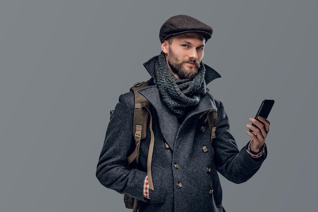 Il ritratto di un viaggiatore con la barba vestito con una giacca e un berretto di lana tiene uno zaino, utilizzando uno smartphone.