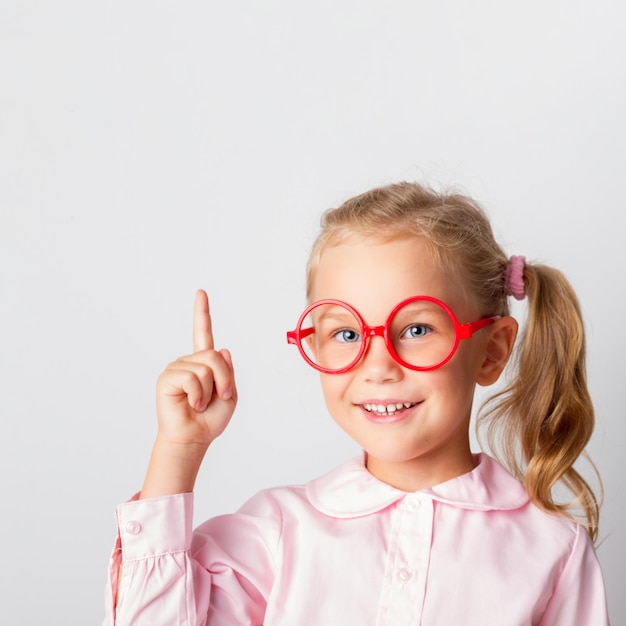 Il ritratto di una bambina intelligente con gli occhiali rossi che tiene un pollice in alto ha un'idea su uno sfondo chiaro