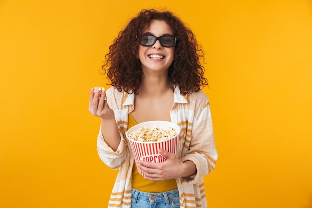 Il ritratto di una bella giovane donna riccia sveglia che posa isolata sulla parete gialla con gli occhiali mangia popcorn guarda film.