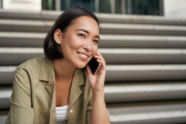 Il ritratto di una bella ragazza asiatica parla al cellulare si siede sulle scale della strada Donna con lo smartphone che sorride effettuando una chiamata
