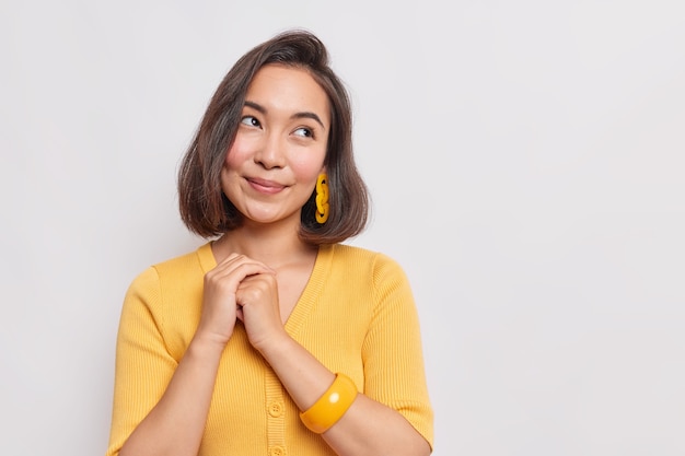 Il ritratto di una bella ragazza asiatica tiene le mani unite distoglie lo sguardo con l'espressione sognante che sogna qualcosa indossa un braccialetto casual giallo sul braccio isolato sul muro bianco