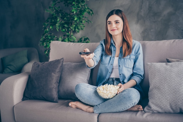 Il ritratto di una bella ragazza positiva si siede sul divano con le gambe incrociate e guarda il film mangia popcorn