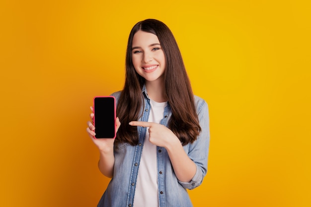 Il ritratto di una bella ragazza sorridente felice tiene lo spazio vuoto del touchscreen del dito diretto del telefono su sfondo giallo