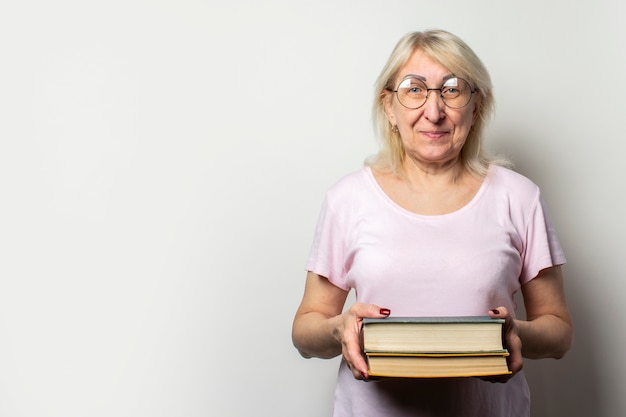 Il ritratto di una donna anziana amichevole con il sorriso in una maglietta e gli occhiali casuali tiene una pila di libri su una parete leggera isolata. Volto emotivo. Concept book club, tempo libero, educazione