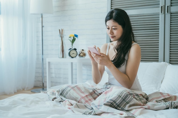 il ritratto di una donna asiatica attraente sorridente è seduto sul letto utilizzando il telefono cellulare dopo essersi alzato la mattina in un accogliente interno della camera da letto di casa con la luce del giorno.