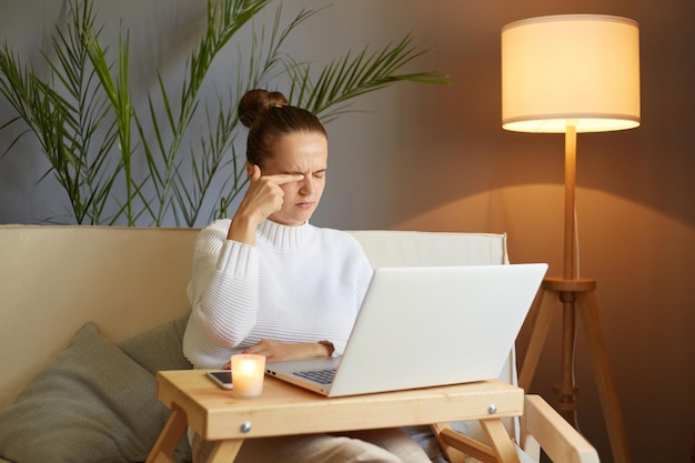 Il ritratto di una donna assonnata triste stanca che indossa un maglione bianco seduto sul divano e lavora su un notebook sente dolore agli occhi strofinandosi gli occhi sentendosi affaticato