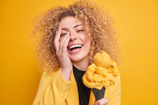 Il ritratto di una donna dai capelli ricci positiva fa sorridere il palmo della faccia e si sente felicemente molto felice di godersi il giorno libero in compagnia di amici divertenti mangia un delizioso gelato ad alto contenuto calorico non si preoccupa della dieta