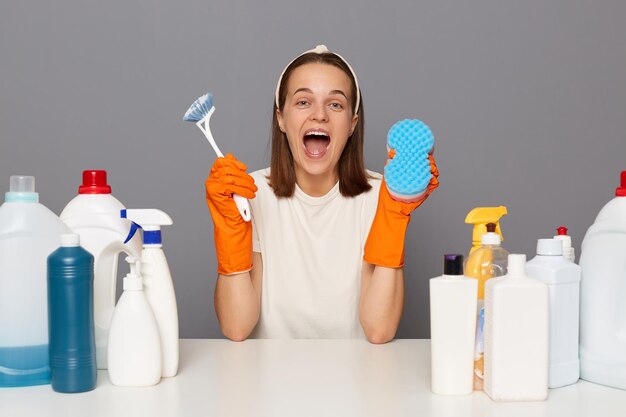 Il ritratto di una donna eccitata e felicissima indossa guanti arancioni con fascia e una maglietta casual si siede sul posto di lavoro utilizza diversi detergenti che tengono pennello e spolverino in pp nelle mani isolate su sfondo grigio