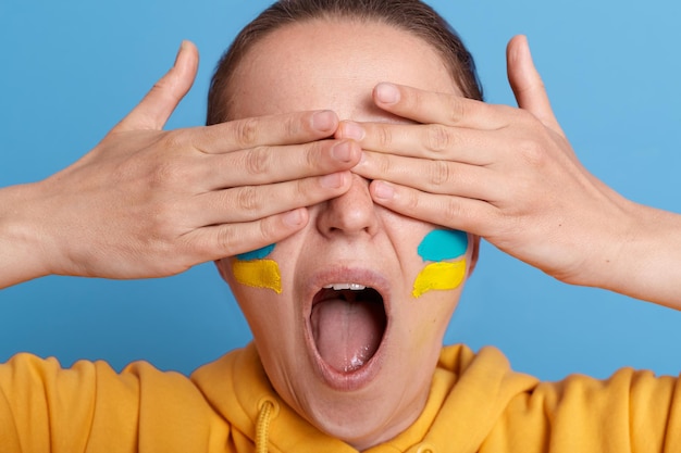 Il ritratto di una donna ucraina che si copre gli occhi con i palmi delle mani urlando forte mantiene la bocca ampiamente aperta essendo spaventoso sente paura a causa della guerra che esprime shock