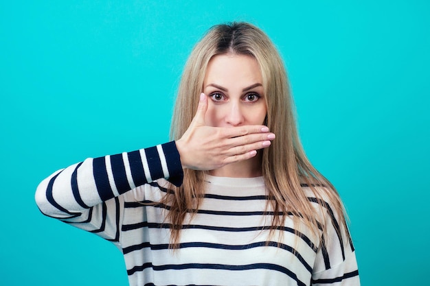 Il ritratto di una giovane donna attraente con il trucco tiene una mano del dito vicino alle labbra in studio su sfondo blu il concetto di segretezza e silenzio