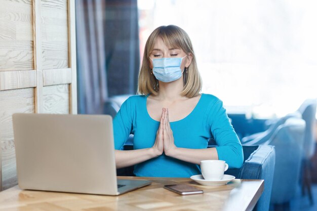 Il ritratto di una giovane donna bionda calma con maschera medica chirurgica in maglietta blu è seduto e lavora sul laptop e cerca di rilassarsi nella posa yoga. Concetto di lavoro al chiuso, medicina e assistenza sanitaria.