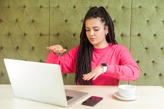 Il ritratto di una giovane donna cieca di concentrazione seria con l'acconciatura di dreadlocks neri in camicetta rosa è seduto al caffè e si copre gli occhi cercando di trovare un laptop con le braccia alzate Stile di vita al chiuso