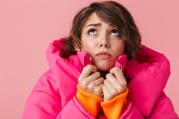 Il ritratto di una giovane donna dispiaciuta con un cappotto caldo si congela e guarda verso l'alto isolato sul rosa