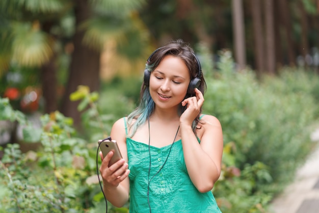 Il ritratto di una giovane donna intelligente in estate copre l'ascolto della musica con le cuffie mentre tiene il telefono cellulare sull'erba attraverso le palme. Concetto di libertà.