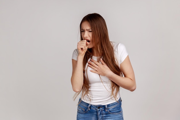 Il ritratto di una giovane donna malata adulta con i capelli scuri ha tosse e dolore ai polmoni, ha sintomi di malattia, indossa una maglietta bianca. Studio indoor girato isolato su sfondo grigio.