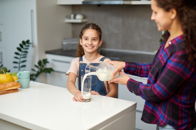 Il ritratto di una mamma versa il latte della figlia carina nel bicchiere per colazione