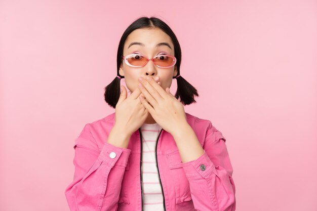 Il ritratto di una ragazza asiatica alla moda scioccata in occhiali da sole chiude gli sguardi della bocca con l'espressione del viso sorpresa si erge su sfondo rosa