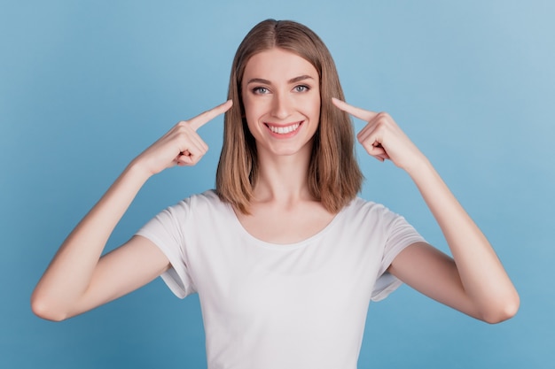 Il ritratto di una ragazza sana e allegra positiva indica un sorriso raggiante con gli occhi delle dita su sfondo blu