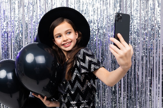 Il ritratto di una ragazza sorridente su una celebrazione di compleanno prende un selfie su uno smartphone fra i palloni neri dell'elio su un brillante