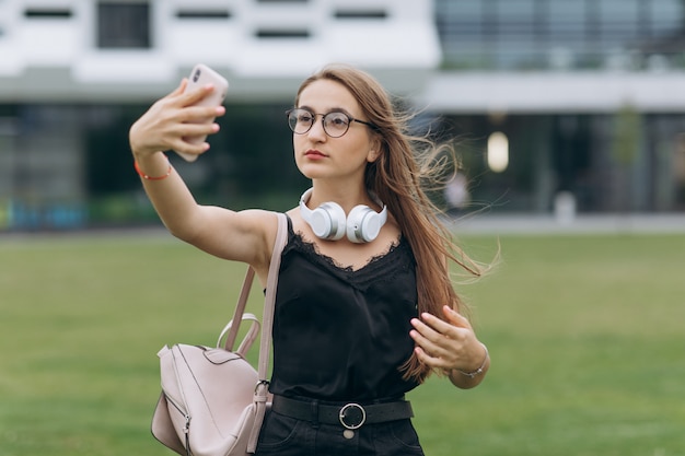 Il ritratto di una signora sorridente felice allegra della scolara del giovane studente con gli occhiali d'uso dei capelli lunghi prende un selfie.