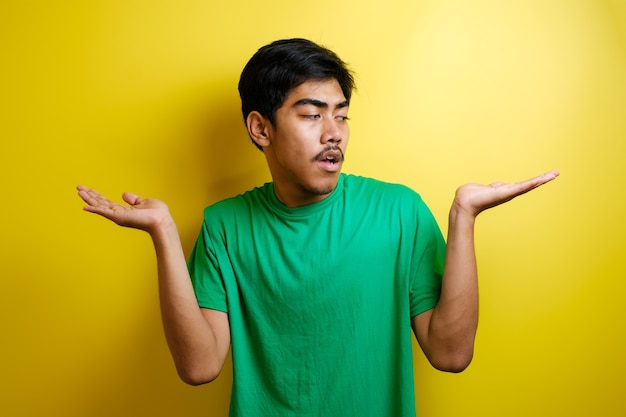 Il ritratto di uno studente universitario asiatico maschio che indossa una maglietta verde mostra le scelte tra i palmi delle mani vuote, il concetto di dubbio su sfondo giallo