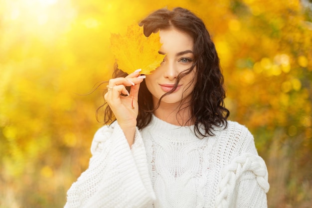 Il ritratto femminile autunnale di una bella donna riccia fresca in un maglione bianco vintage alla moda cammina nel parco e copre l'occhio con una foglia d'acero caduta gialla nel parco dorato al tramonto