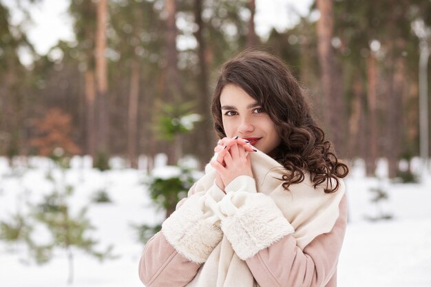 Il ritratto invernale di una donna bruna contenta indossa una sciarpa e un cappotto caldo che cammina nella foresta