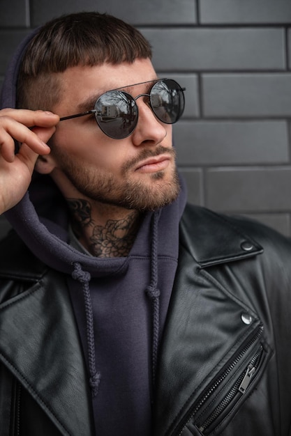 Il ritratto maschile urbano di un bell'uomo brutale e serio hipster in abito nero alla moda con giacca rock in pelle e felpa con cappuccio indossa occhiali da sole alla moda su uno sfondo di mattoni neri